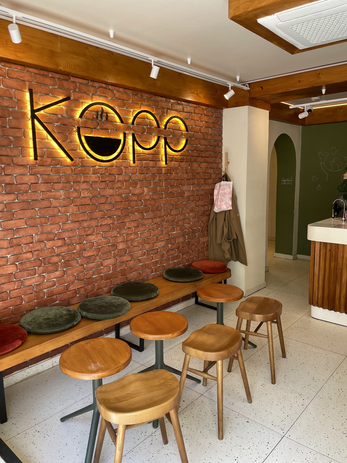 Logo-ul KOPP retroiluminat pe peretele de cărămidă, deasupra băncii cu perne colorate