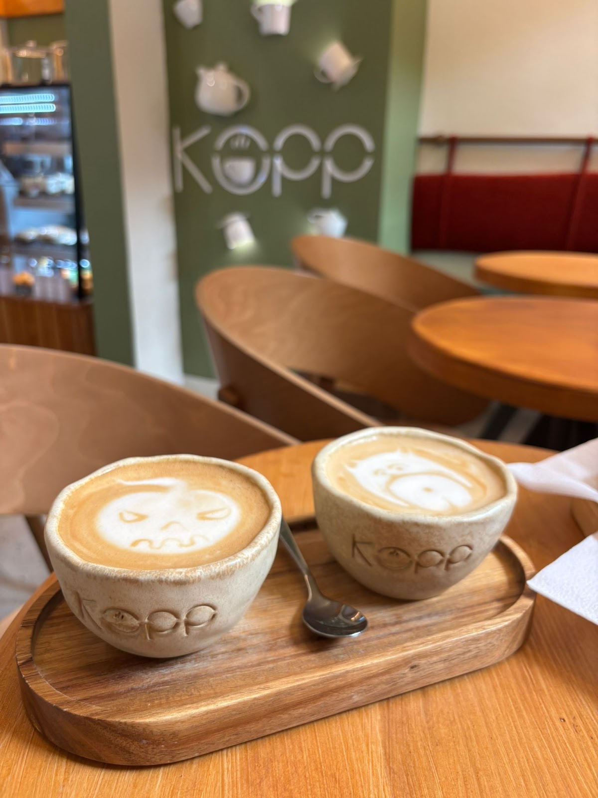 Două cappuccino cu latte art în cești KOPP, sub logo-ul de pe peretele verde