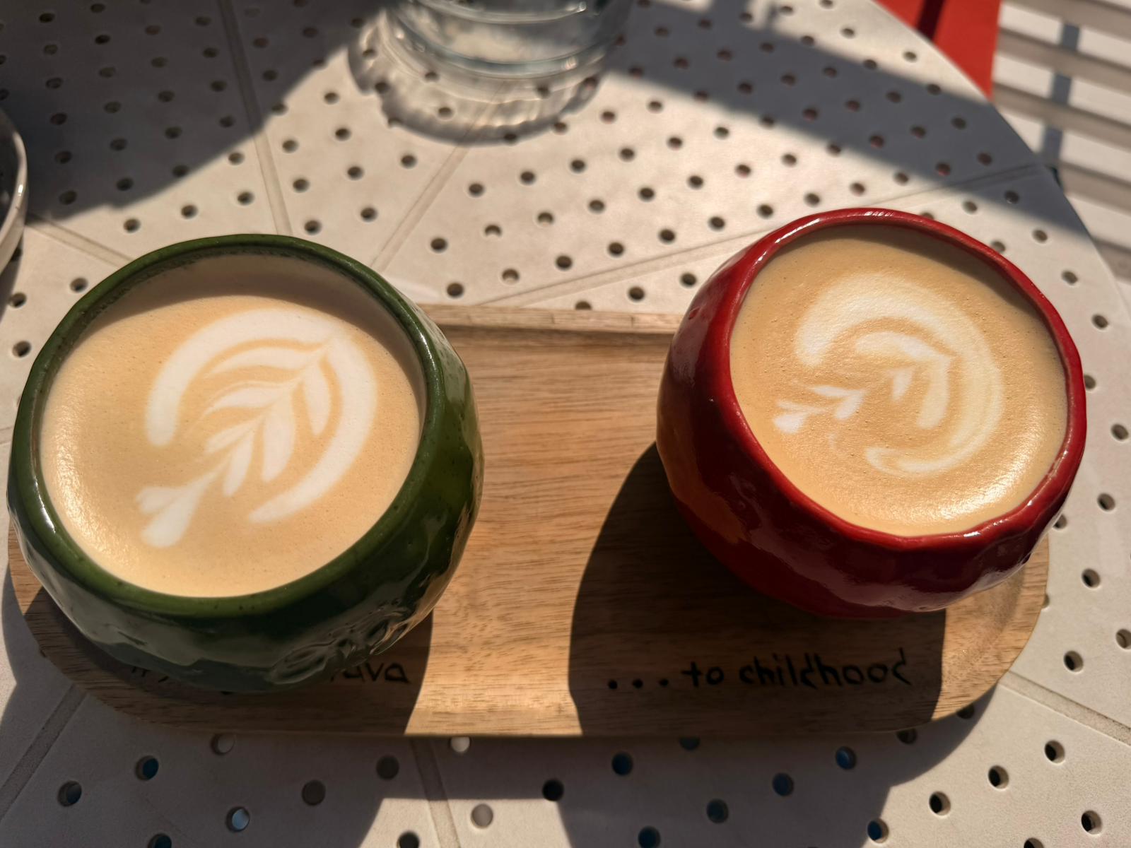 Două cafele cu latte art în cești verde și roșu, pe o tavă cu mesajul to childhood